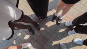 A group of individuals stands in a circle on a sunlit paved surface. Only their feet and legs are visible, showcasing a variety of stylish footwear, including black heels, loafers, and white sneakers. The ground consists of gray and brown bricks forming a pattern.